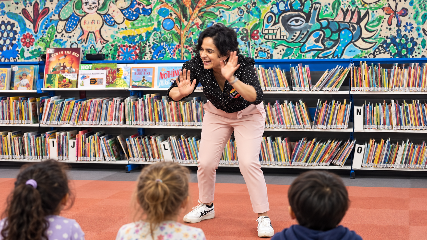 Rohini Vij telling a story with animated hand gestures to children at St Kilda Library, with bookshelves and a colourful mural behind her
