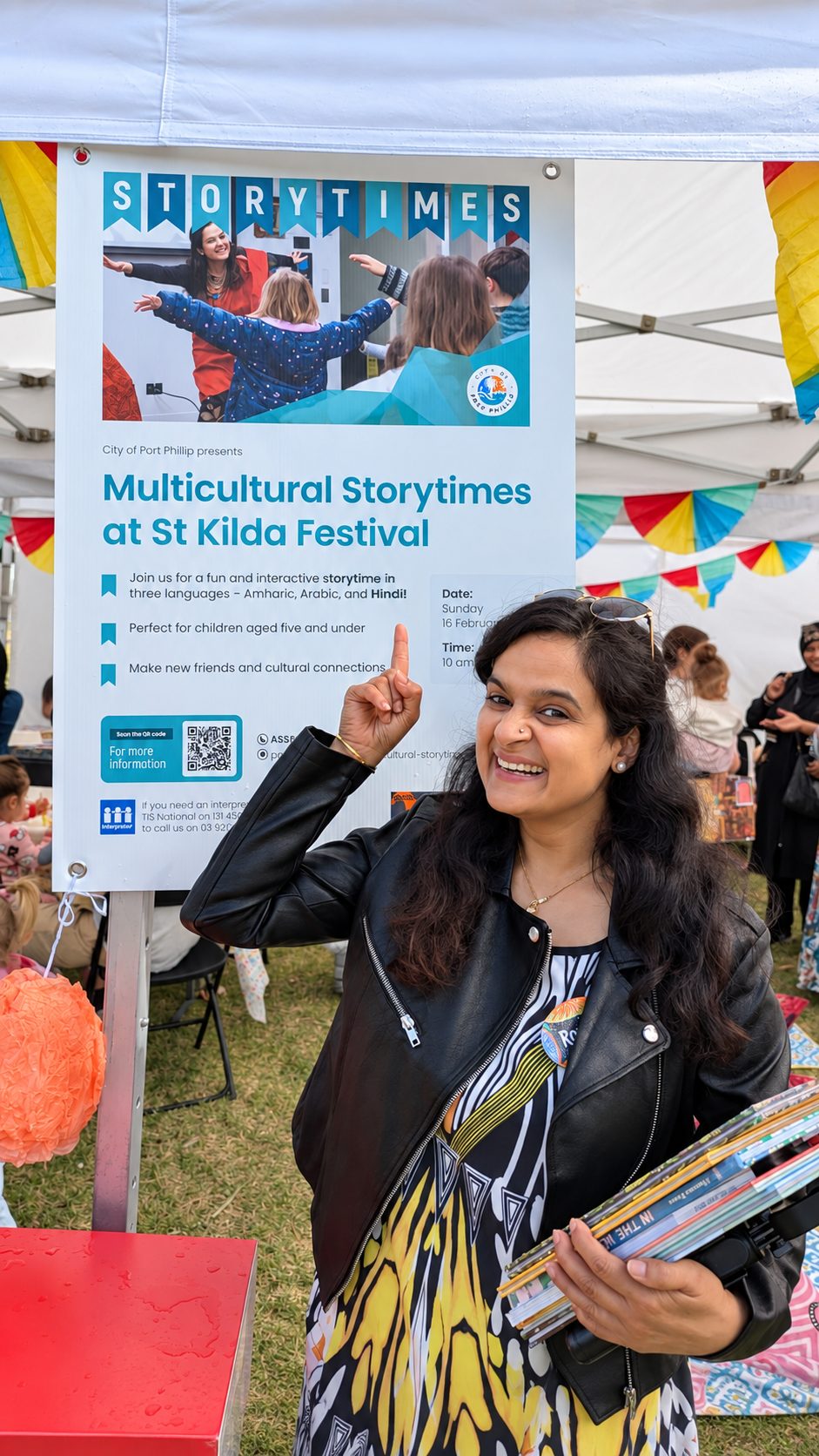 Rohini Vij holding children’s books and pointing to the Multicultural Storytimes sign at St Kilda Festival
